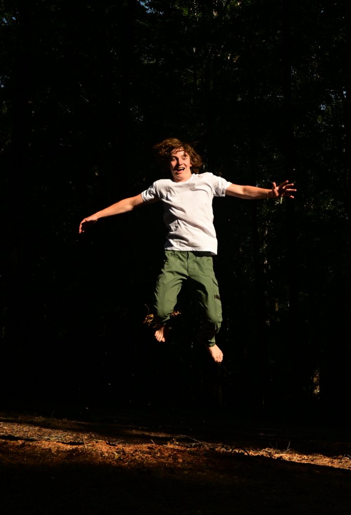 Greyson mid-jump in a clearing, arms out, barefoot, lit against dark pines