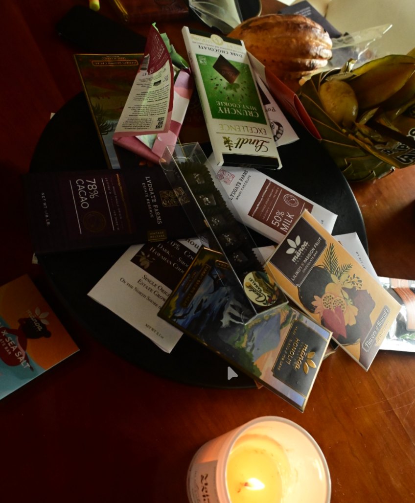 A chocolate tasting laid out on a dark round tray: a dozen bars fanned across it, a cacao pod and leaves behind, a lit candle in the foreground